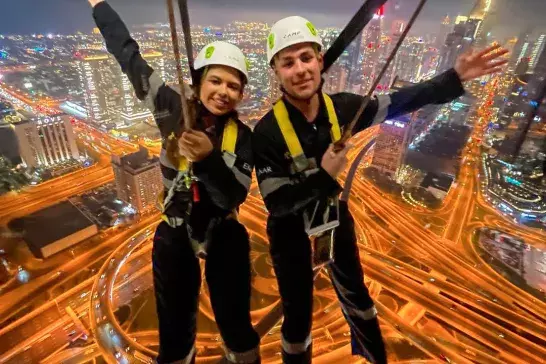 Two individuals wearing safety harnesses and helmets are suspended above a brightly lit cityscape at night. One of them is Travel Advisor Cassidy Royea, enjoying an adventurous activity, possibly zip-lining or bungee jumping.