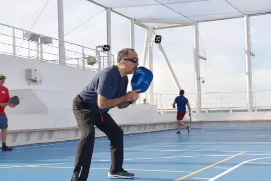 People play pickleball on a blue court aboard a ship, with railings and open sky in the background.
