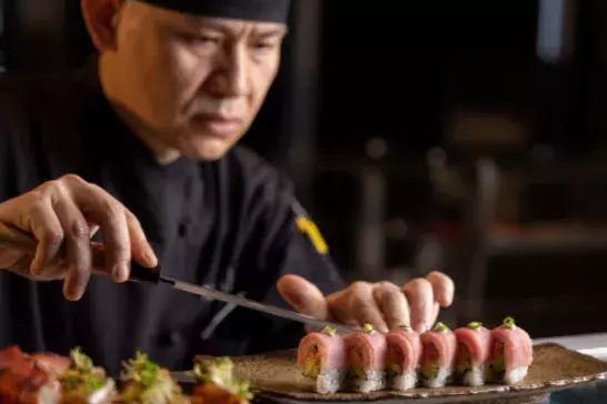 A chef in a black uniform prepares sushi, arranging pieces on a rectangular plate.