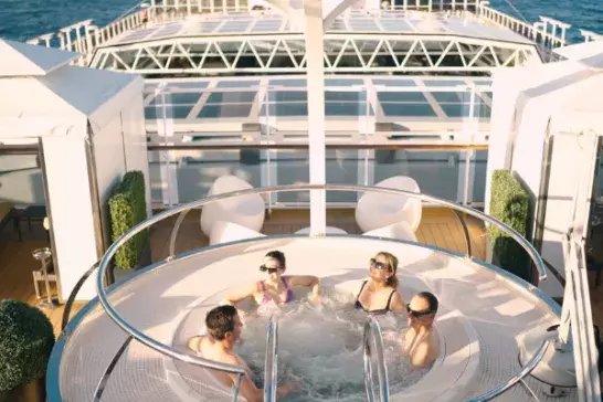 Four people relax in a circular hot tub on a cruise ship deck with ocean views in the background.