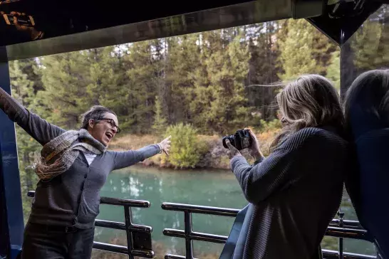 Two people on a balcony overlooking a river and forest; one poses while the other takes a photo.