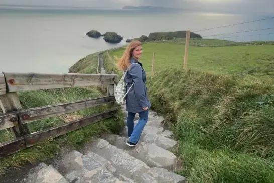 Travel Advisor Shayna walks down a stone staircase toward a coastal path in Ireland, with grassy fields and ocean views under an overcast sky