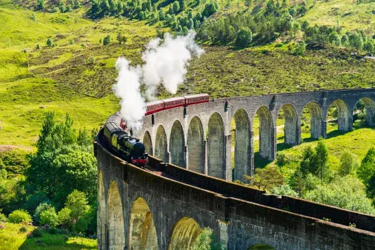 Steam train crossing a curved viaduct in a green hilly landscape.