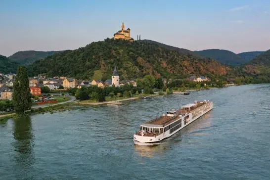 A large white river cruise ship sails along a wide river, passing by a picturesque village with numerous houses and buildings. The village is nestled at the base of a lush green hill, atop which stands an impressive castle. The surrounding landscape features rolling hills and dense forests under a clear blue sky.