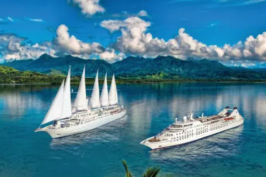 Two large cruise ships sail on calm, clear water surrounded by lush green mountains and a partly cloudy sky. Tropical palm trees frame the foreground. One ship features multiple sails, while the other is a modern cruise liner.