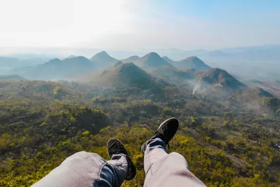 Legs in jeans and black shoes overlooking misty green mountains from a high viewpoint.