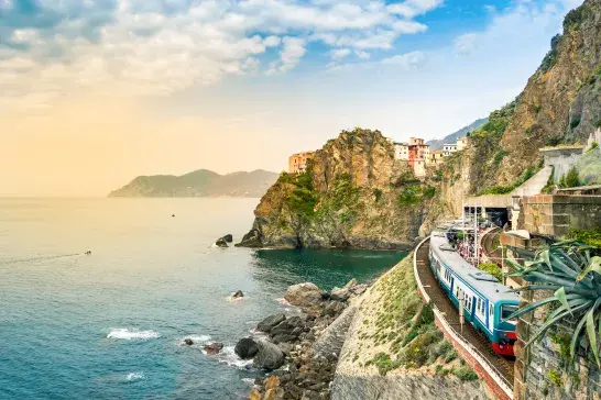 A train traveling along a coastal railway track on a steep rocky cliff overlooking the blue sea, with colorful buildings perched on the cliffside under a partly cloudy sky.