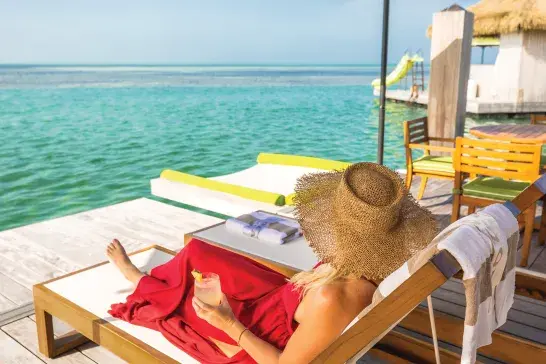 Person relaxing on a lounge chair by turquoise ocean under a green umbrella at an overwater deck.