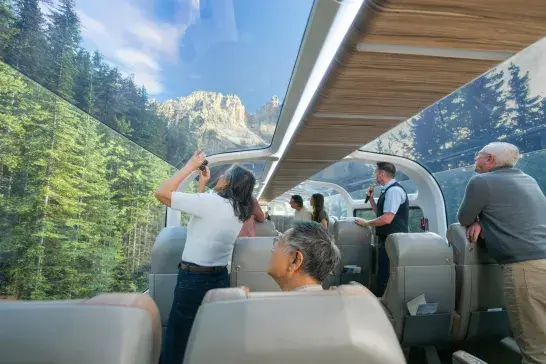 Scenic train interior with glass dome windows showing stunning mountain peaks and dense forest, passengers taking photos and enjoying panoramic views.