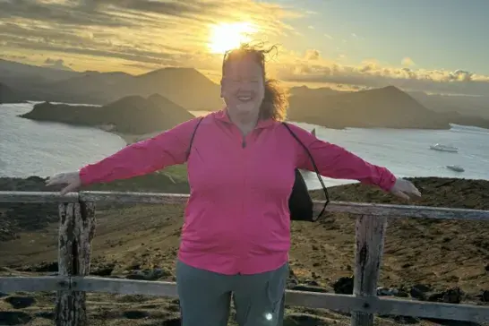 Trish standing with arms outstretched against a wooden railing, overlooking a coastal landscape with island hills and ocean below at sunset.