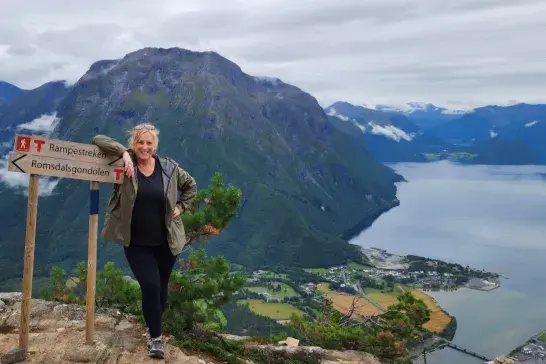 Trisha Hall posing next to a Romsdalsgondolen trail sign on a rocky mountain viewpoint, with fjords, steep green mountains, and a shoreline village below.