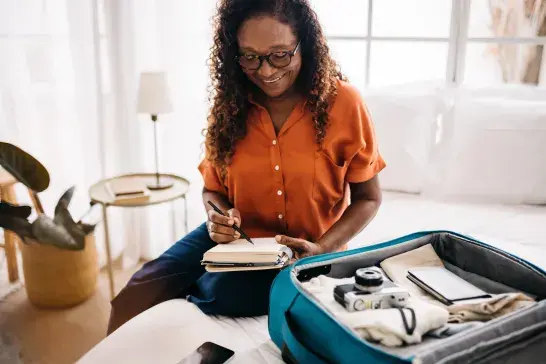 Person sitting on a bed writing in a notebook beside an open suitcase filled with travel items.