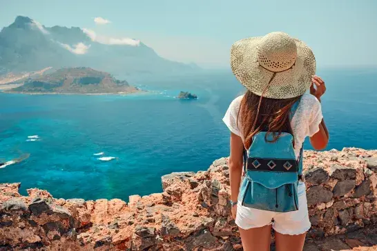 A person wearing a sunhat, white shirt, and a small blue backpack stands on a rocky viewpoint overlooking a bright blue sea with distant mountains and a small island in the background.