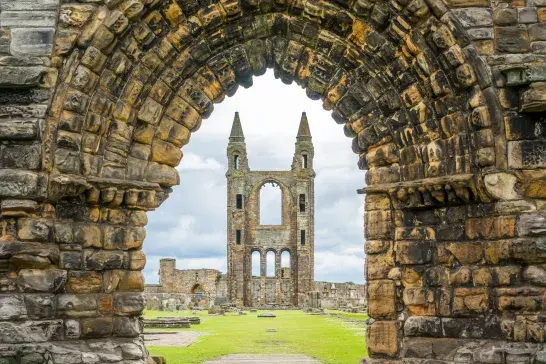 Stone archway framing the ruins of St Andrews Cathedral in St Andrews, Scotland, with a grassy courtyard and the cathedral’s central tower visible beyond.