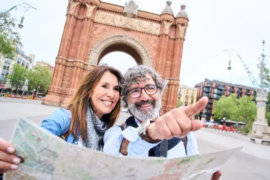 Two people stand in front of a large red-brick archway, holding a map as one of them points ahead. Buildings, trees, and a wide pedestrian walkway surround the landmark.