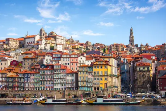 Colorful historic buildings with red-tiled roofs line the hillside along the Douro River in Porto, Portugal, with traditional boats moored on the water in the foreground and church towers rising above the city under a blue sky.