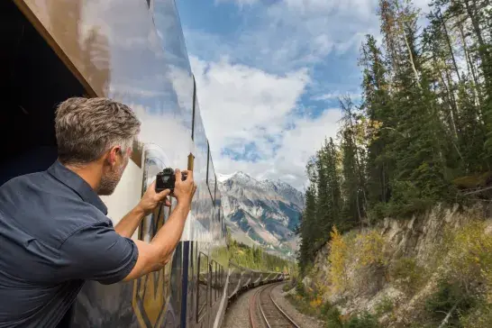 A traveller leans out of a Rocky Mountaineer train to take a photo as the train curves through forested mountains in the Canadian Rockies