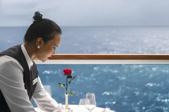 A staff member setting a dinner table on the cruise ship balcony overlooking the water