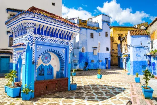 Blue and white buildings in a Moroccan street, featuring intricate architectural details, tiled rooftops, and a decorative fountain in the foreground under a bright blue sky.