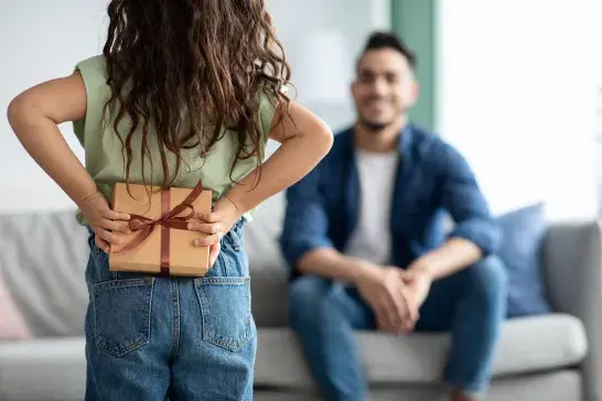 Child holding a wrapped gift behind their back while standing in front of an adult seated on a couch