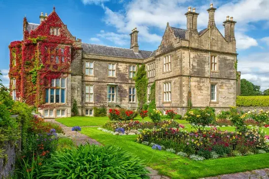 Historic stone manor house covered in red and green ivy, surrounded by colourful flower gardens, manicured lawns, and stone pathways under a blue sky.