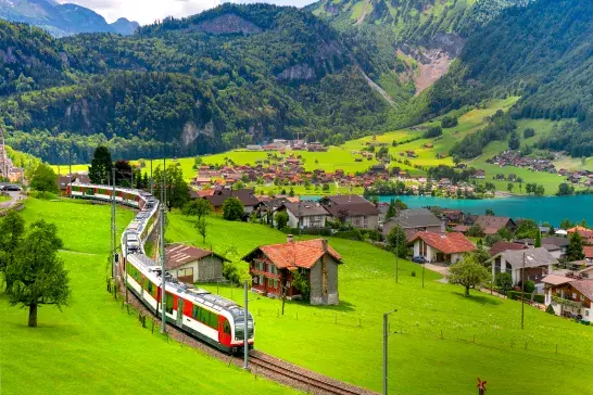 A passenger train winds through lush green countryside past scattered homes and farmland, with a turquoise lake, a village, and forested mountains rising in the background.