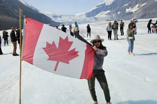 A person stands on a glacier holding a large Canadian flag while surrounded by other visitors. Snow-capped mountains and an expansive ice field stretch into the distance under a clear blue sky. The flag fills much of the foreground, with the glacier and mountain landscape providing a dramatic backdrop.