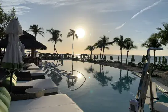 Infinity pool overlooking a tropical beach at sunrise, with palm trees, loungers and ocean views.
