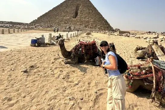 Kailyn standing beside resting camels in front of the Great Pyramid of Giza on a sunny day, with desert sand and historic stone structures in the background.