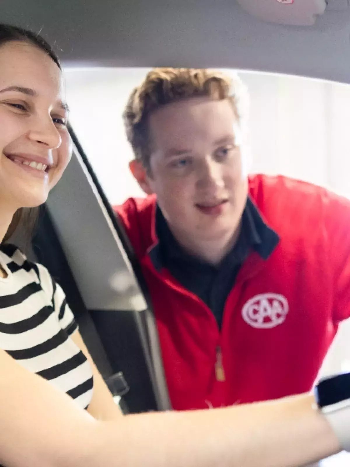 Young female in a stripe tshirt in the driver seat of a car touching the navigation screen with a CAA worker looking through her window asissting