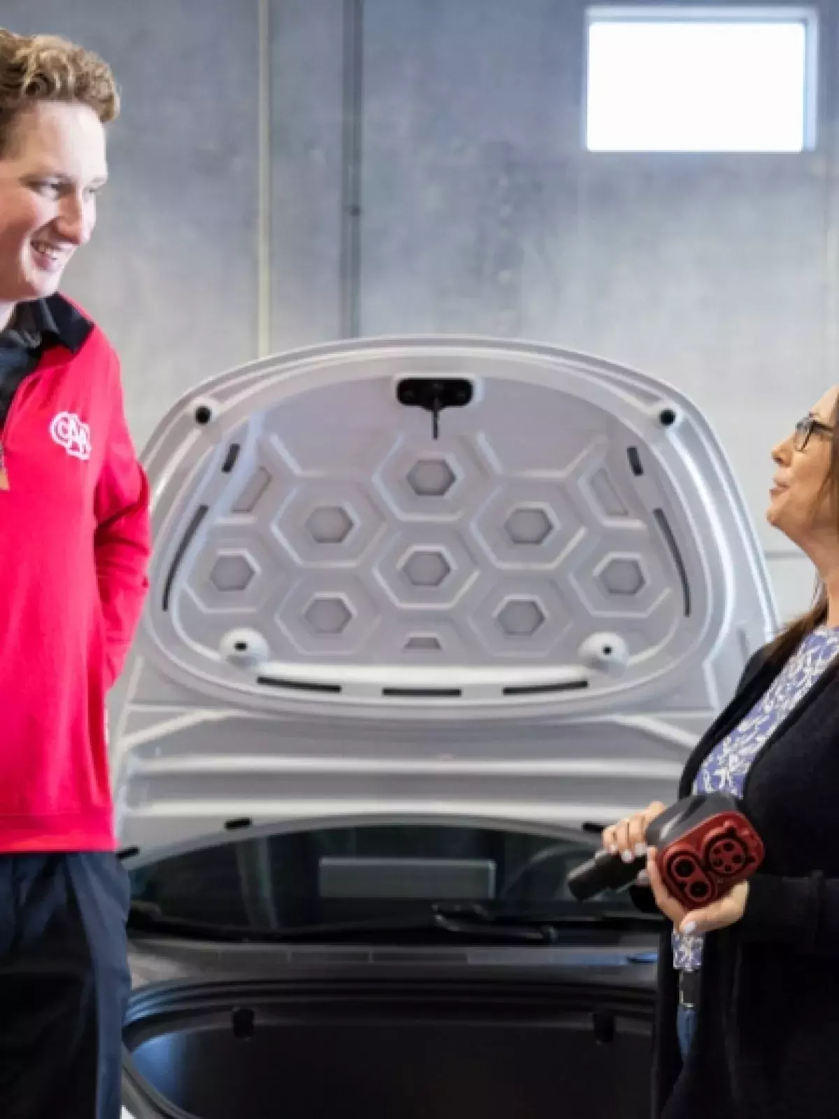 A man in a red CAA sweater looking at a woman holding an electric vehicle charger, both standing in front of a car with its hood up