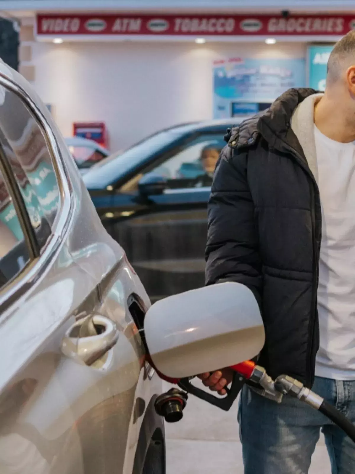 Man in black winter jacket, white tshirt and jeans fueling his silver SUV at a gas station smiling looking at the pump 