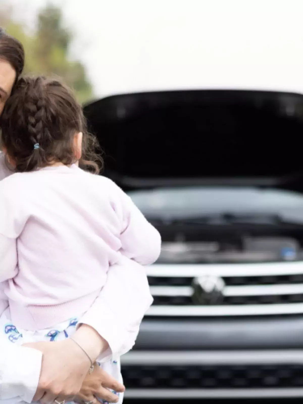 Young mother wearing a white button down shirt holding a small child who is looking back at their vehicle that has the hood open that is being serviced by a roadside technician in neon reflective gear
