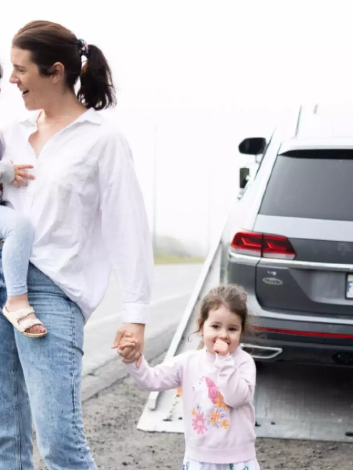young caucasian mother holding one child in her arms and holding another childs hand on the shoulder of a highway. Pictures behind her is a Volkswagen SUV being secured on a tow truck by a roadside assistance worker in neon saefty gear