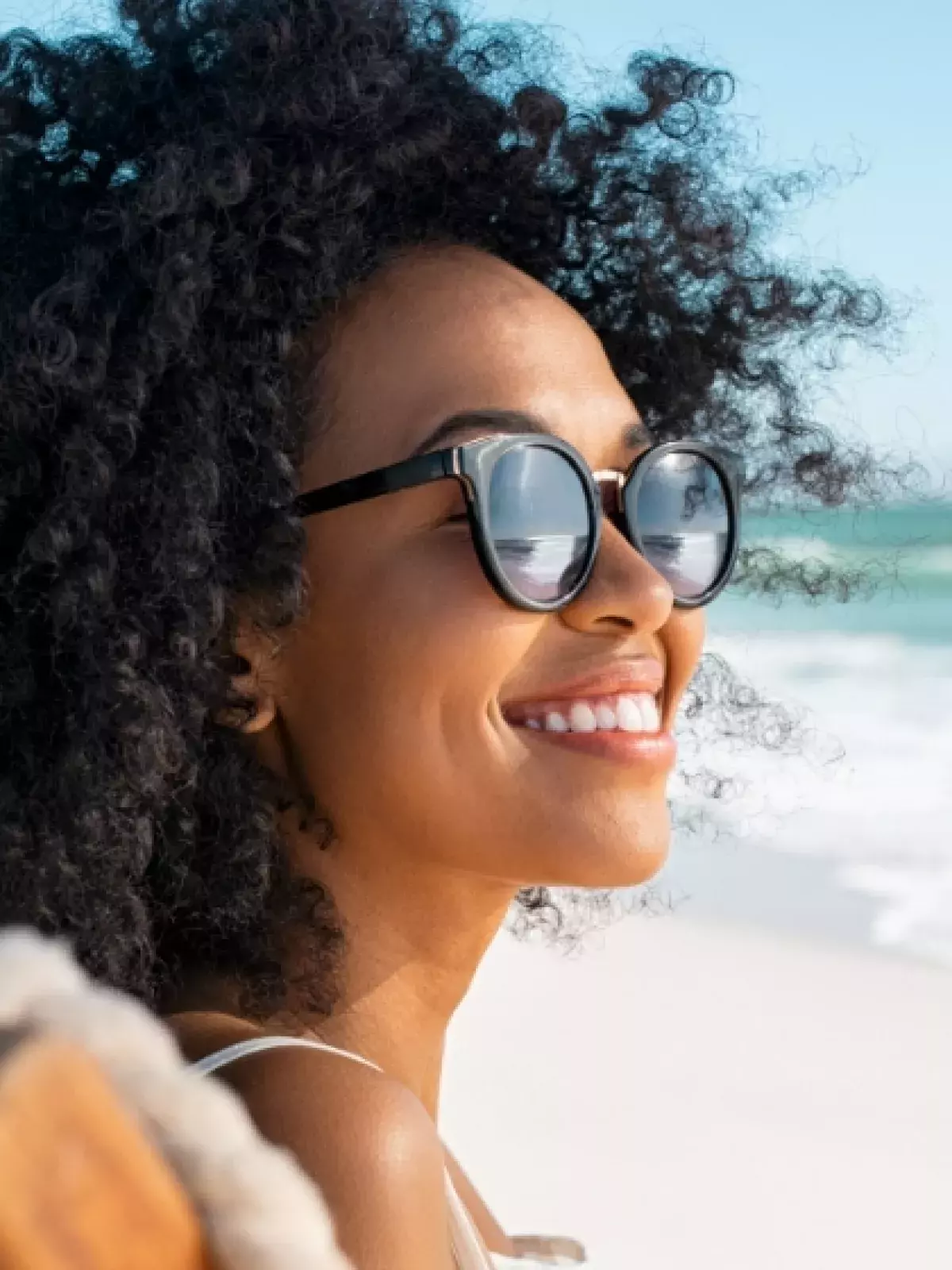 close up shot of a woman smiling with curly hair wearing black sunglasses sitting on a beach chair at the beach 