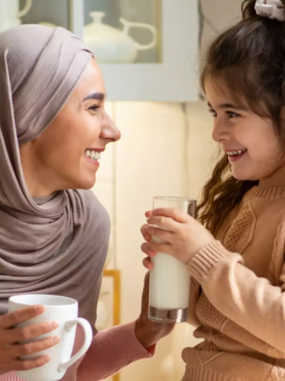 Young mother wearing a pink long sleeve shirt and a grey hijab holding a white coffee cup smiling at a young girl wearing a camel sweater holding a glass of milk in their kitchen