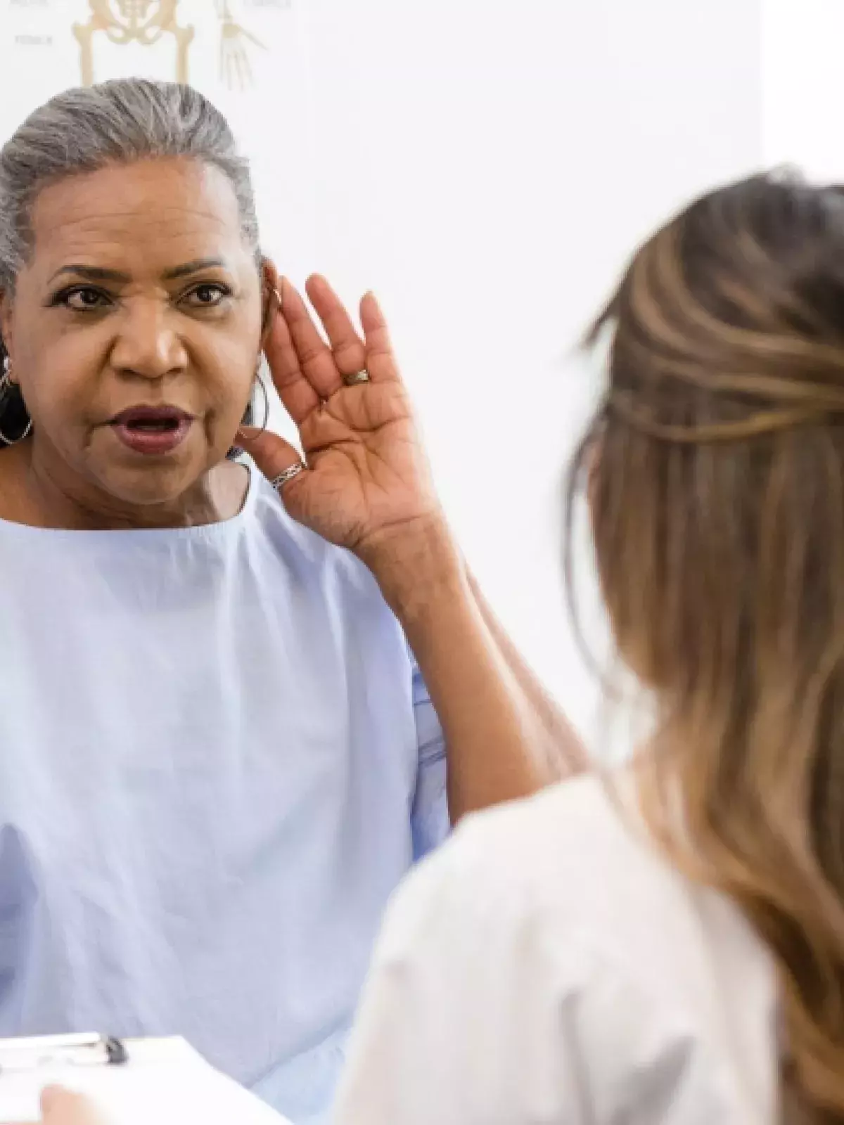 Lady in a doctors office in a blue dressing gown holding up a hand to her ear to a doctor with a clipboard