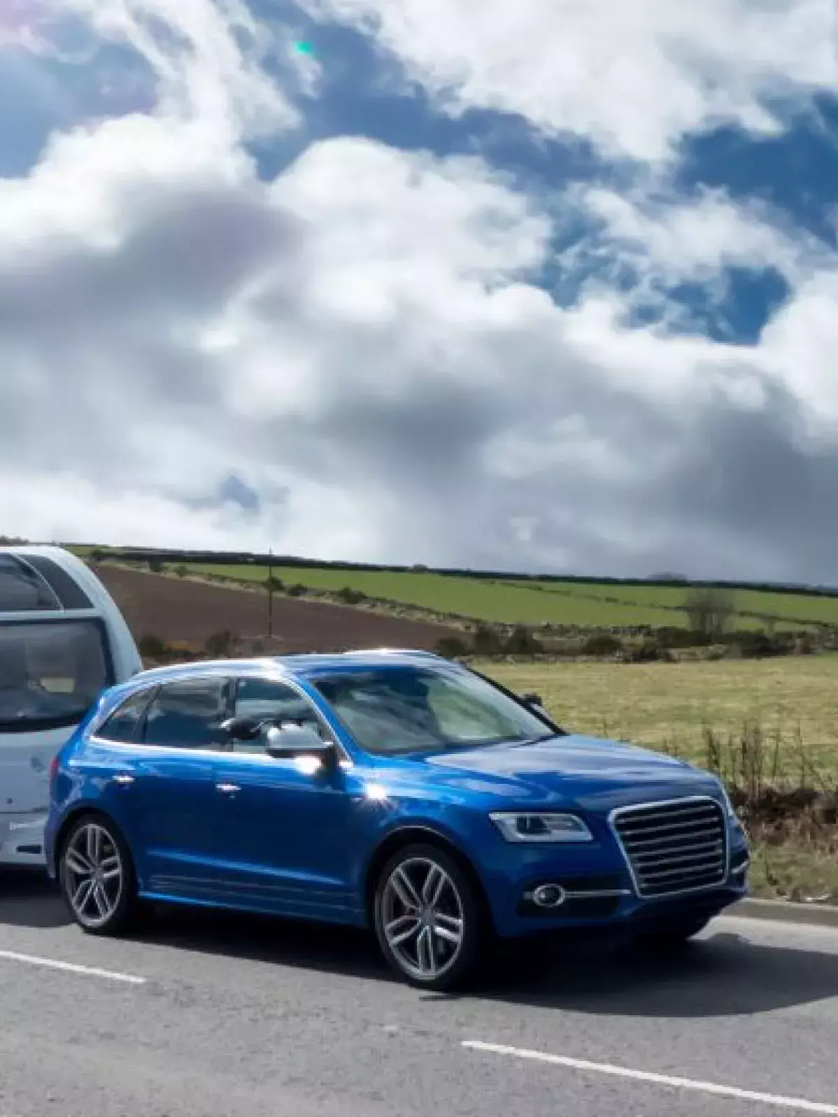 Blue SUV is towing a family camper down a highway surrounded by grassy fields on a sunny day. 