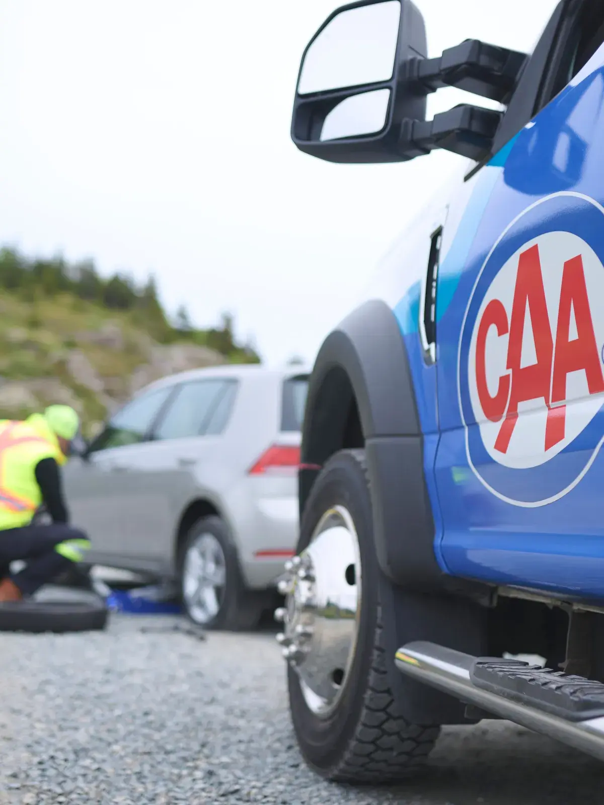 A CAA roadside service vehicle is parked next to a car receiving assistance. The technician, wearing a safety vest, is inspecting the car's front tire with tools on the ground.