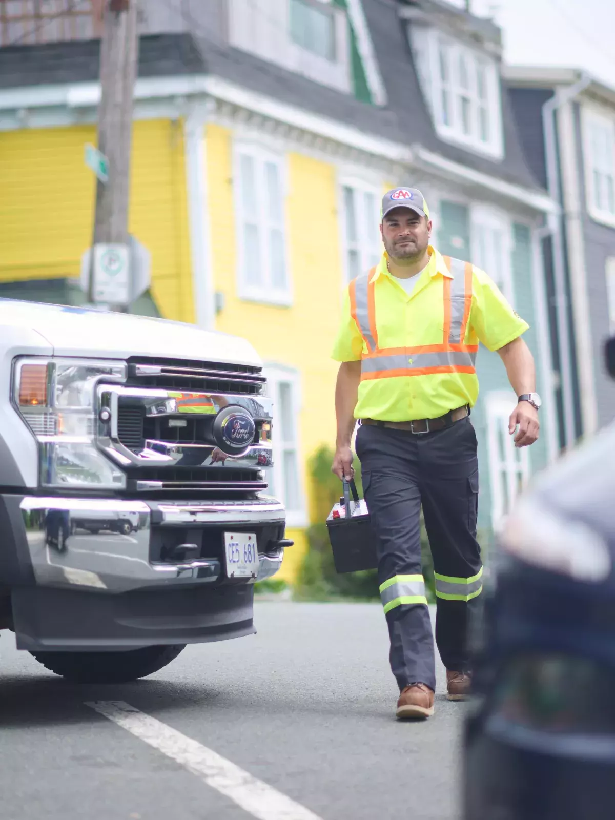 A roadside assistance worker in a high-visibility yellow shirt and cap walks toward a vehicle with an open hood. A service truck with "Member Service" written on the door is parked nearby. Colorful houses line the street in the background.