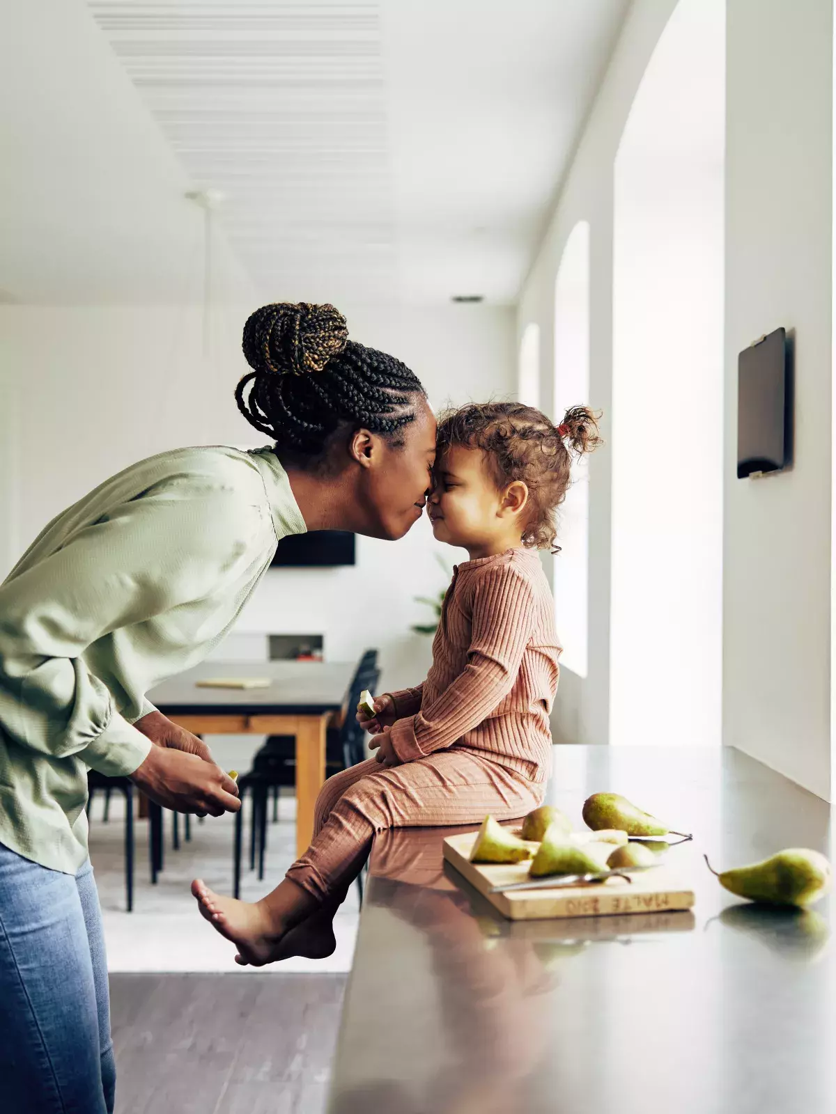 A mother gently kisses her young child on the forehead as the child sits on a kitchen counter. 