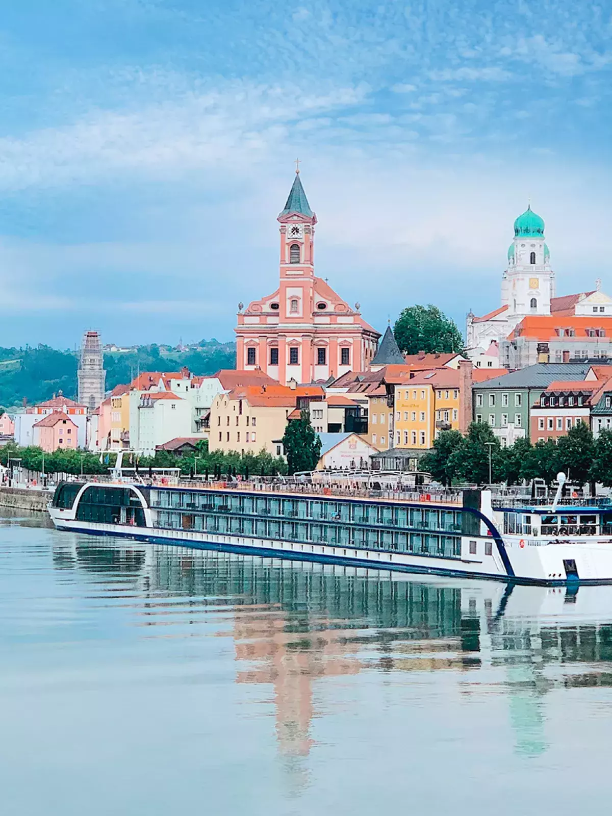 A riverside European town with colorful buildings, a church steeple, and an AmaWaterways river cruise ship docked along the calm water.