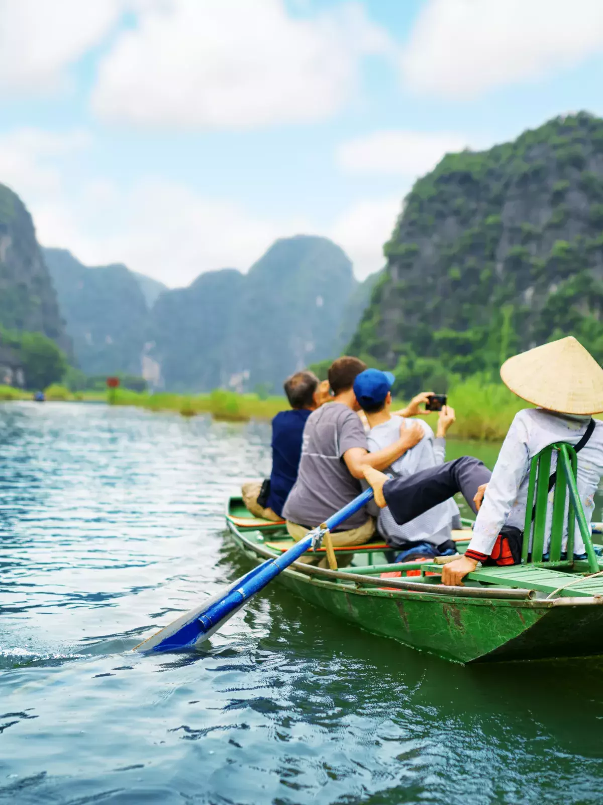 Four people in a green boat on a calm river, surrounded by lush green mountains. One rows with blue oars, another wears a conical hat, and two take photos.