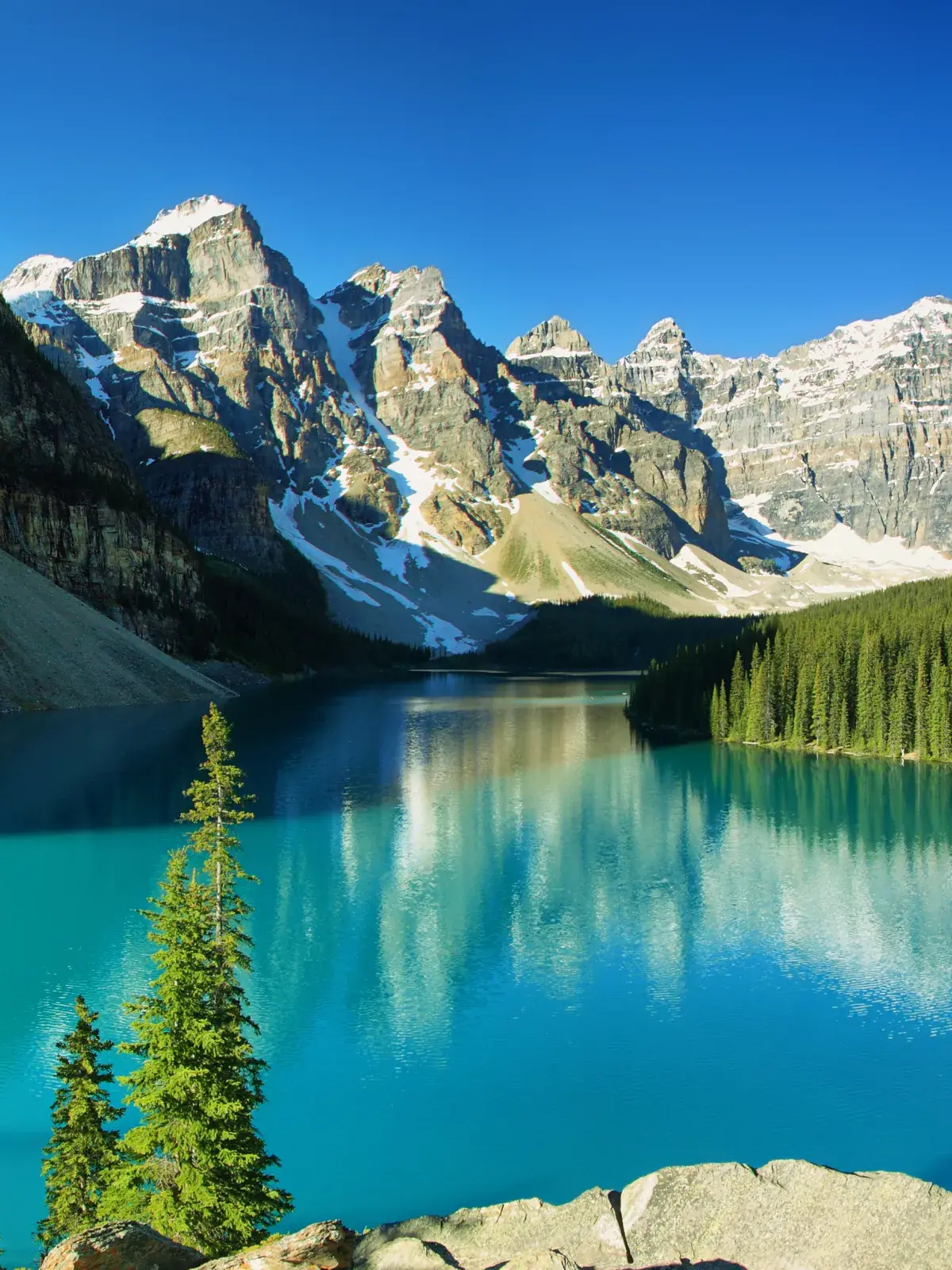 A serene turquoise lake surrounded by dense evergreen forests and towering snow-capped mountains under a clear blue sky.