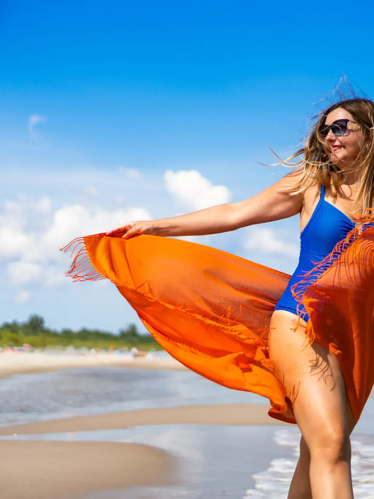 A person wearing a blue swimsuit stands on a sandy beach near the ocean, holding an orange shawl that flows in the wind. The sky is clear and blue with some white clouds, and there are green trees in the background along the shoreline.
