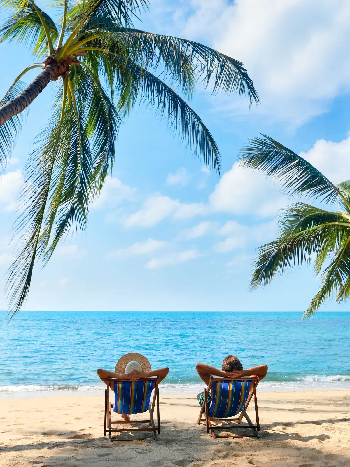 Two people lounge on beach chairs under palm trees by the sea, enjoying a sunny tropical day.