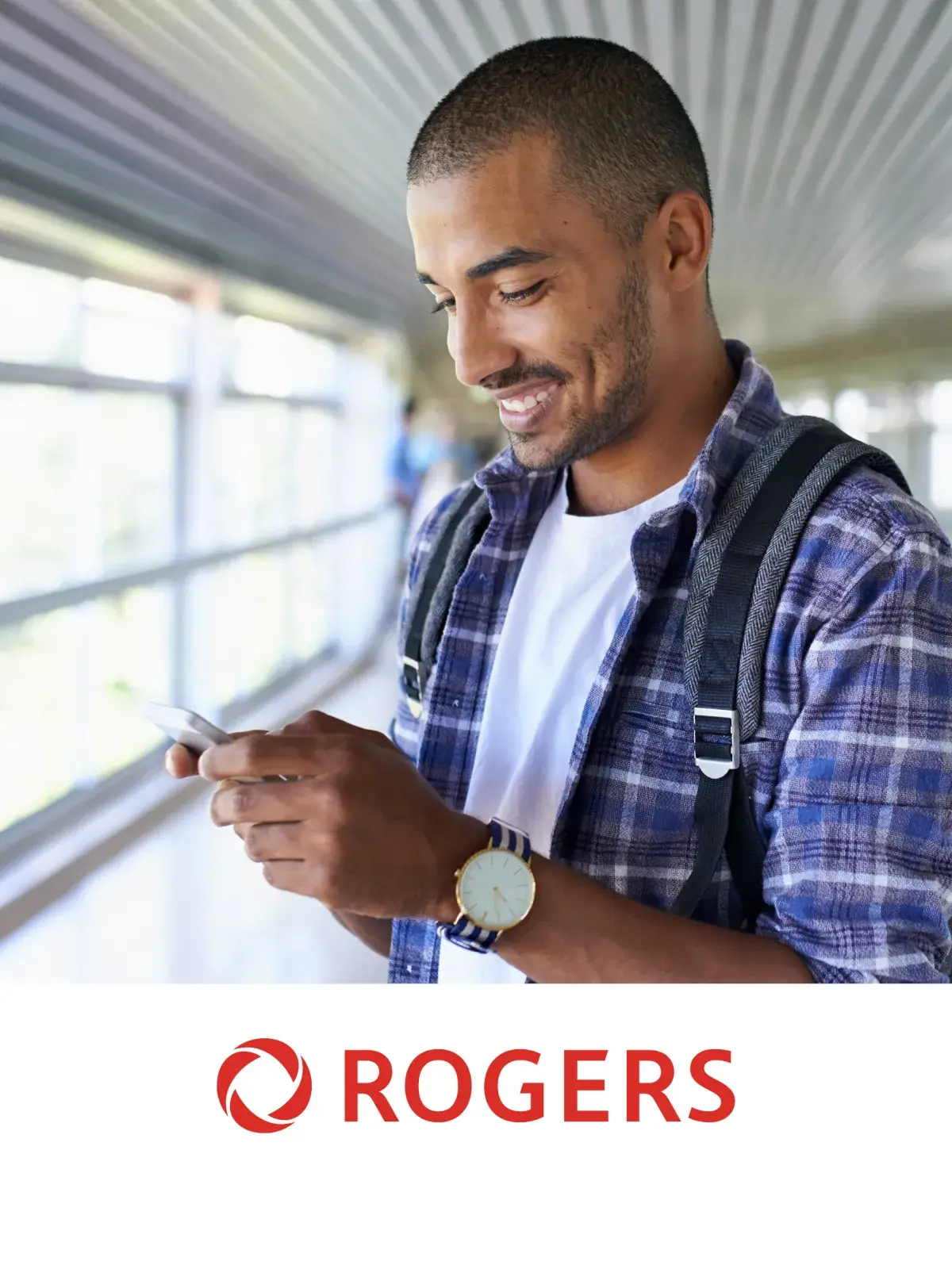 Man in plaid shirt with backpack looking at phone in a bright hallway.