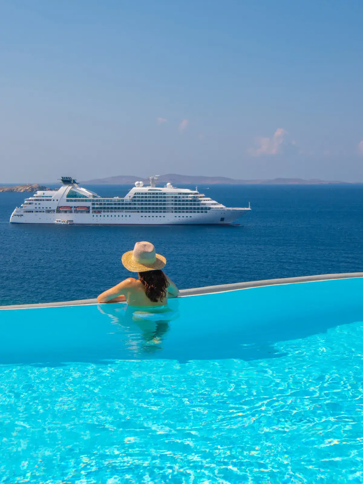 Person relaxing in an infinity pool overlooking a calm blue ocean with a large white cruise ship in the distance under a clear sky.