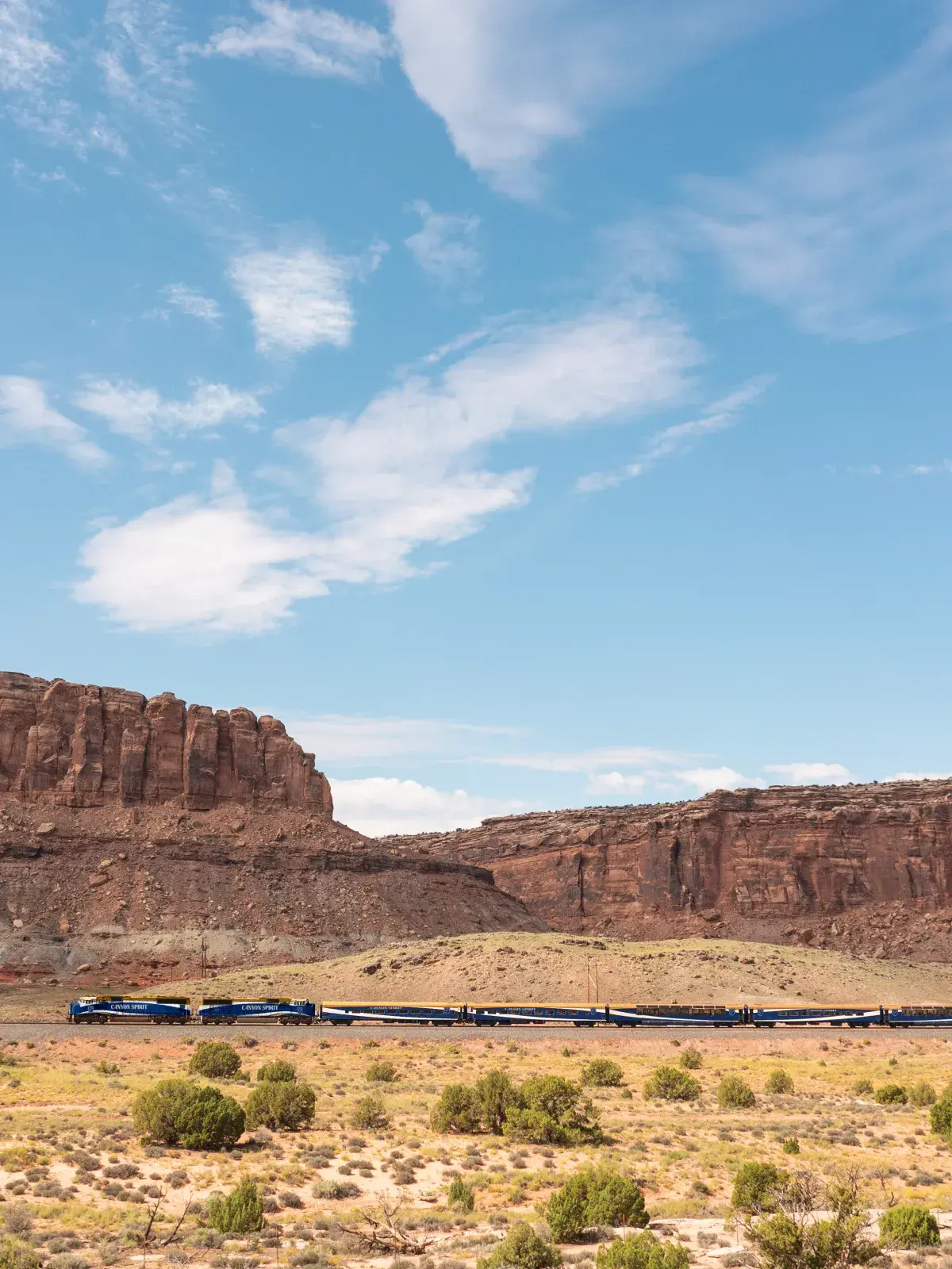 A long passenger train travels across a wide desert landscape with rugged red rock cliffs and a bright blue sky in the background.