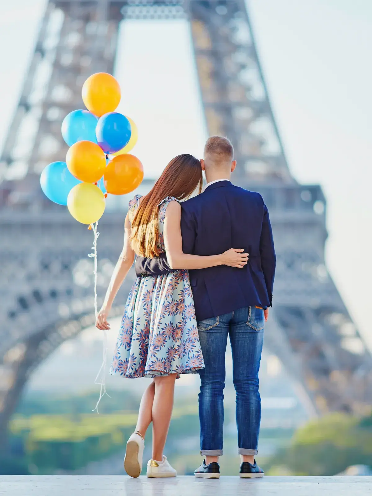 Two people walking away from the camera with arms around each other, holding a cluster of colorful balloons, with the Eiffel Tower blurred in the background on a bright day.